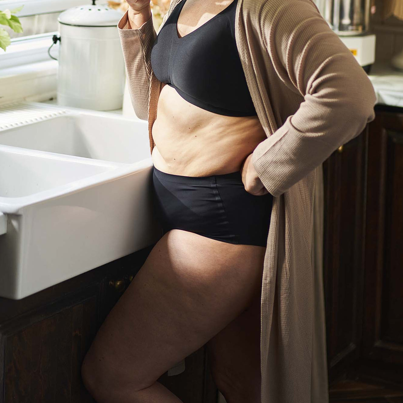 Woman standing in a kitchen holding a mug, wearing a beige cardigan over a black sports bra and shorts.