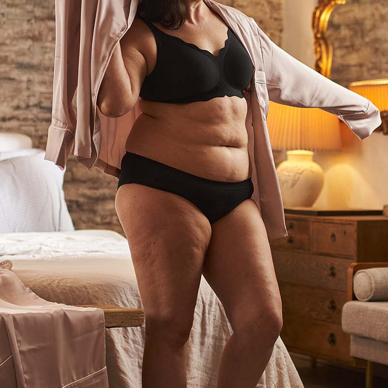 Woman in black lingerie standing in a bedroom with a mirror and lamp in the background.
