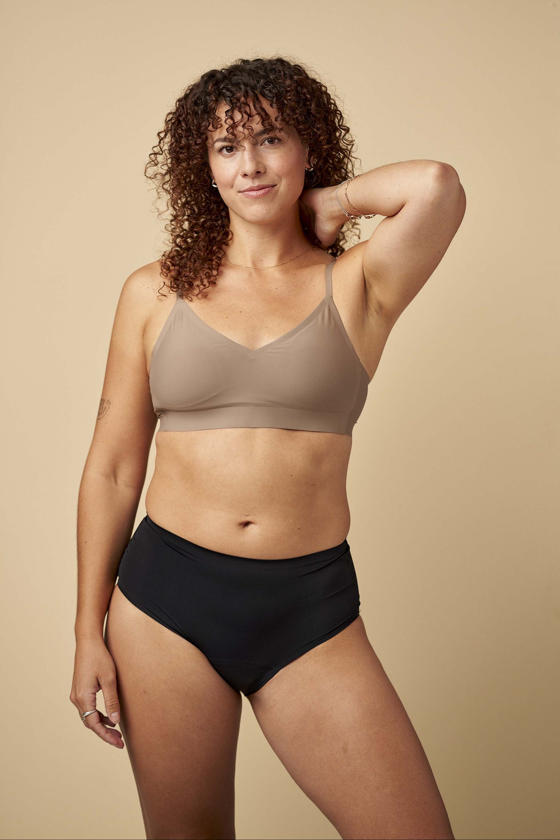 Woman wearing a beige sports bra and black high-waisted underwear against a beige background