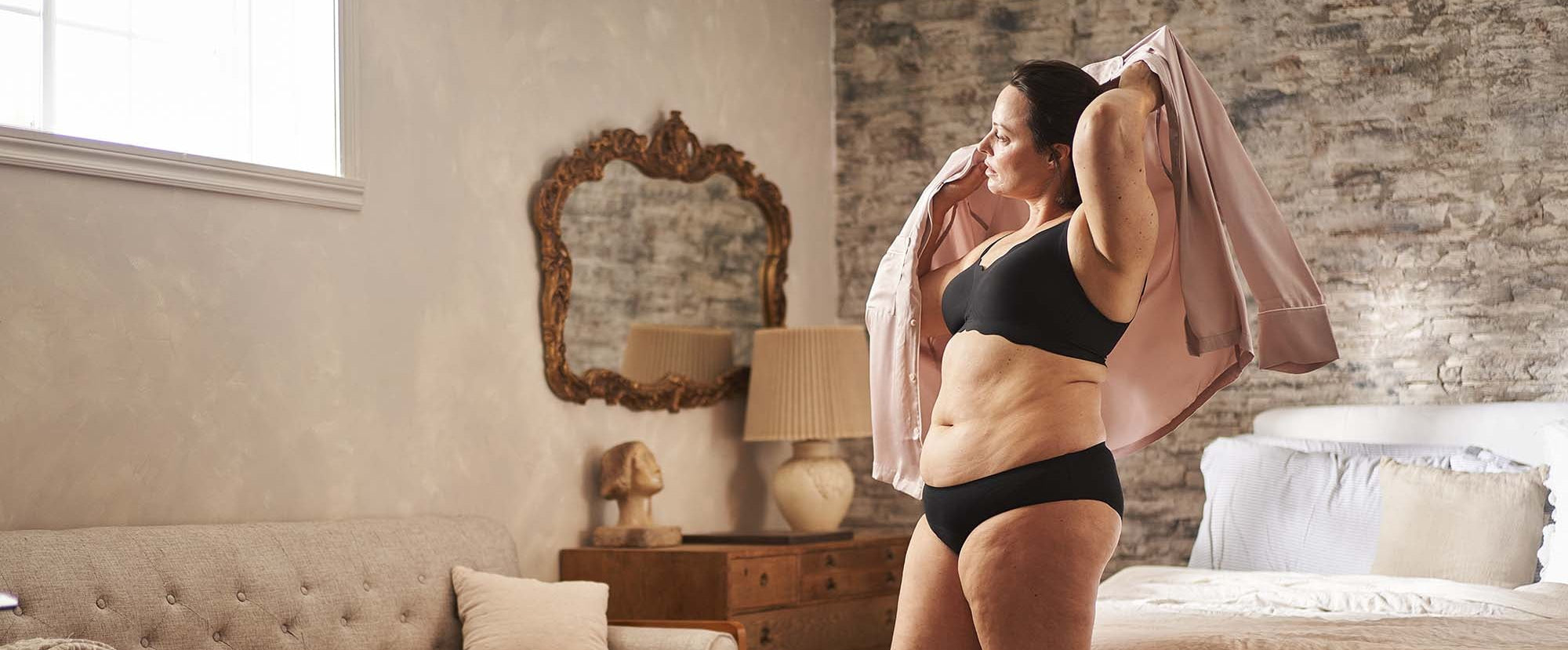 Woman in black lingerie holding a pink bathrobe in a bedroom setting.